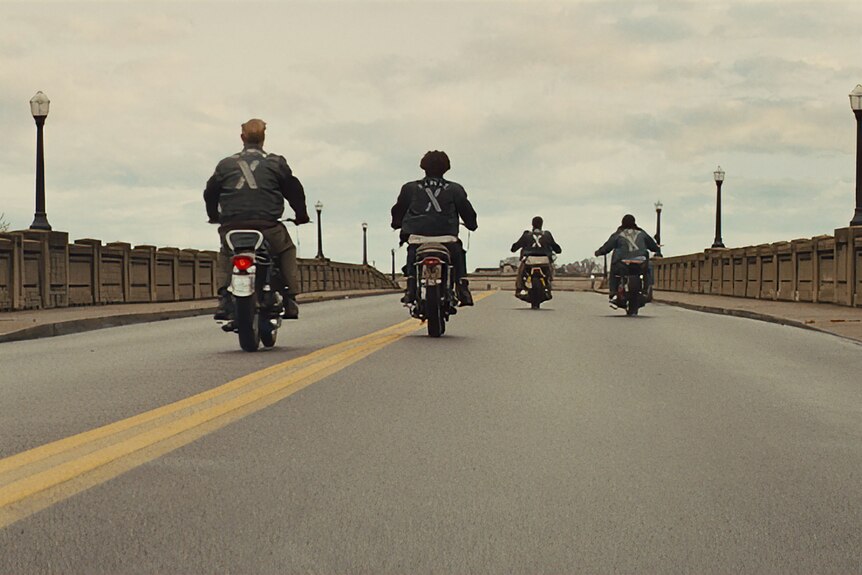 Four motorcyclists ride along a road, pictured from behind.