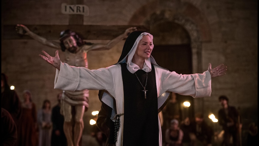 White woman in a nun's habit stands before Christian cross with arms outstretched