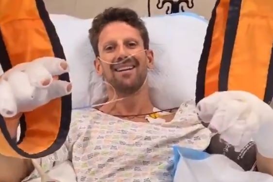 Romain Grosjean smiles while holding up his hands, which are both covered in bangages