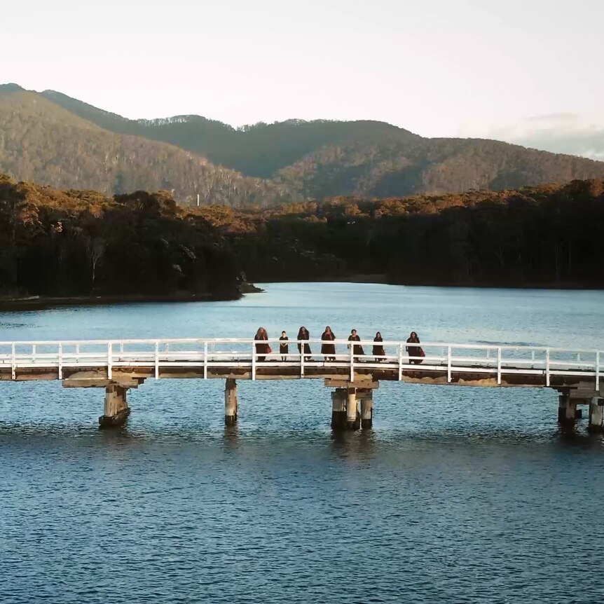 Wide drone shot of a choir standing on a wooden bridge with Gulaga mountain behind them