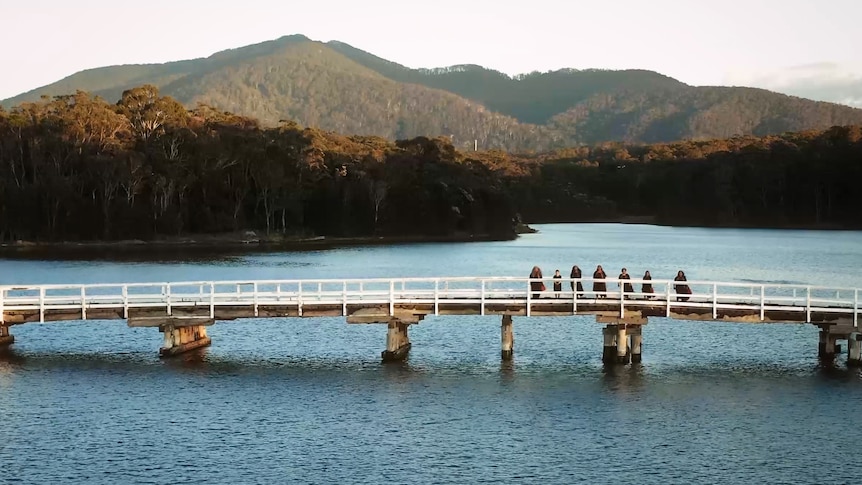 Wide drone shot of a choir standing on a wooden bridge with Gulaga mountain behind them