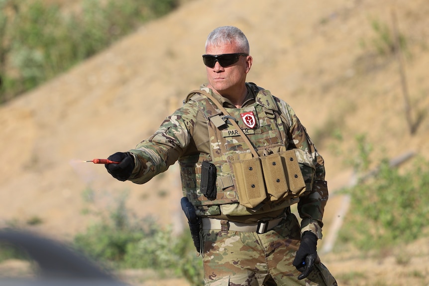 A middle aged man in battle fatigues holds a smoke grenade out in his gloved right hand.