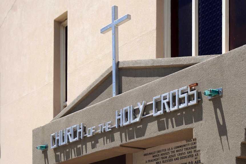 A metal cross above a sign that says "Church of the Holy Cross" on a church building, close up.