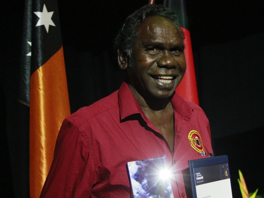 Man looks at camera and holds trophy, standing in front of NT flag and Aboriginal flag