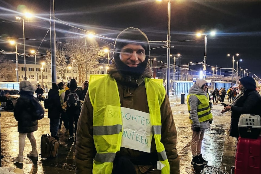 A man stands in a street at night wearing a balaclava and a high-vis vest.