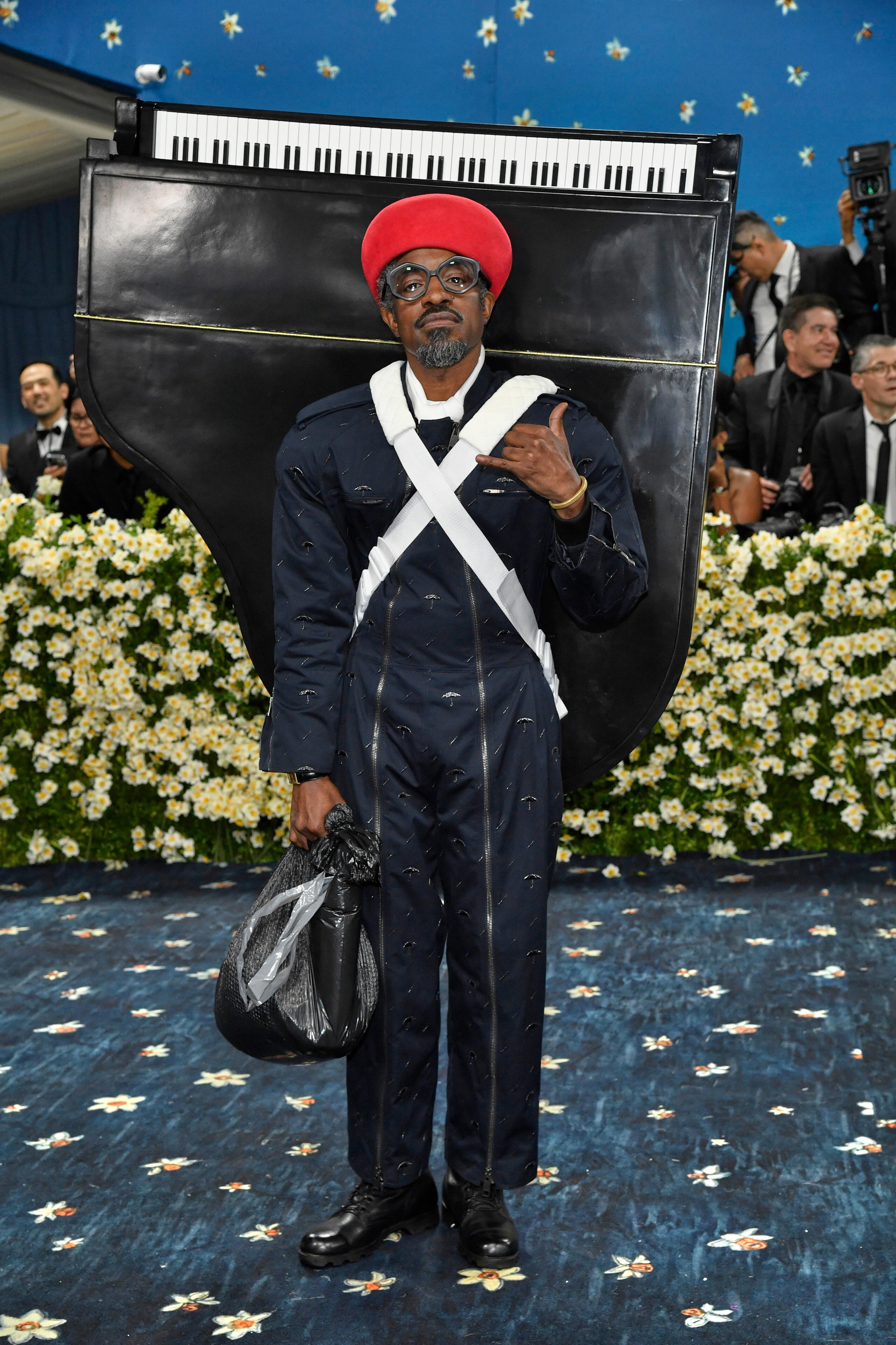 Andre 3000 wearing a prop piano strapped to his back, a red hat and a dark suit