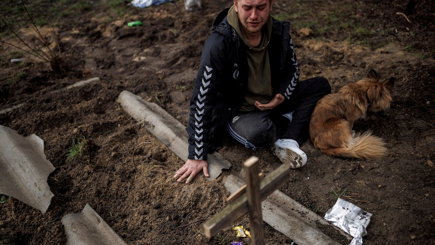 A man sits next to a grave with a dog. 