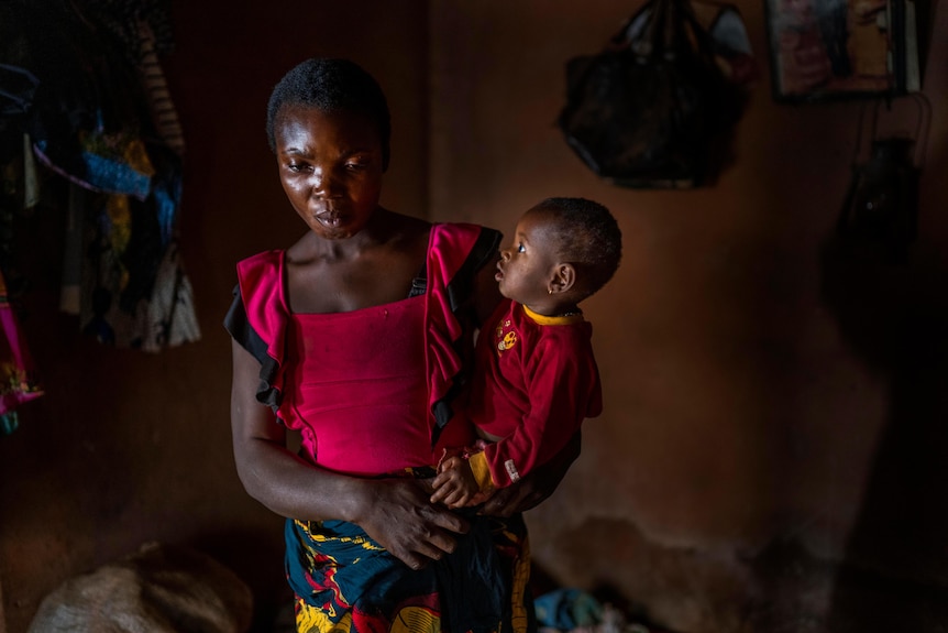 A young mother and African woman holds her young child on her hip while looking to the floor.  
