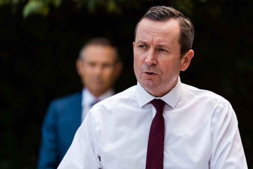 Man in shirt and tie, head and shoulders shot with another man blurred in the background