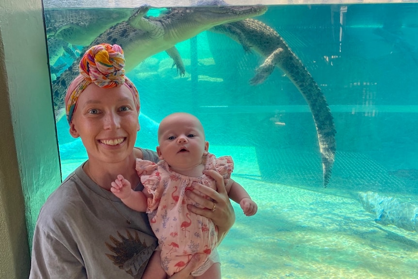 Sarah wears a scarf on her hairless head and holds her baby girl in front of a glass crocodile enclosure