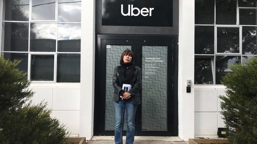 Uber driver Deb standing at a protest outside the company's Melbourne office with sign in the background.