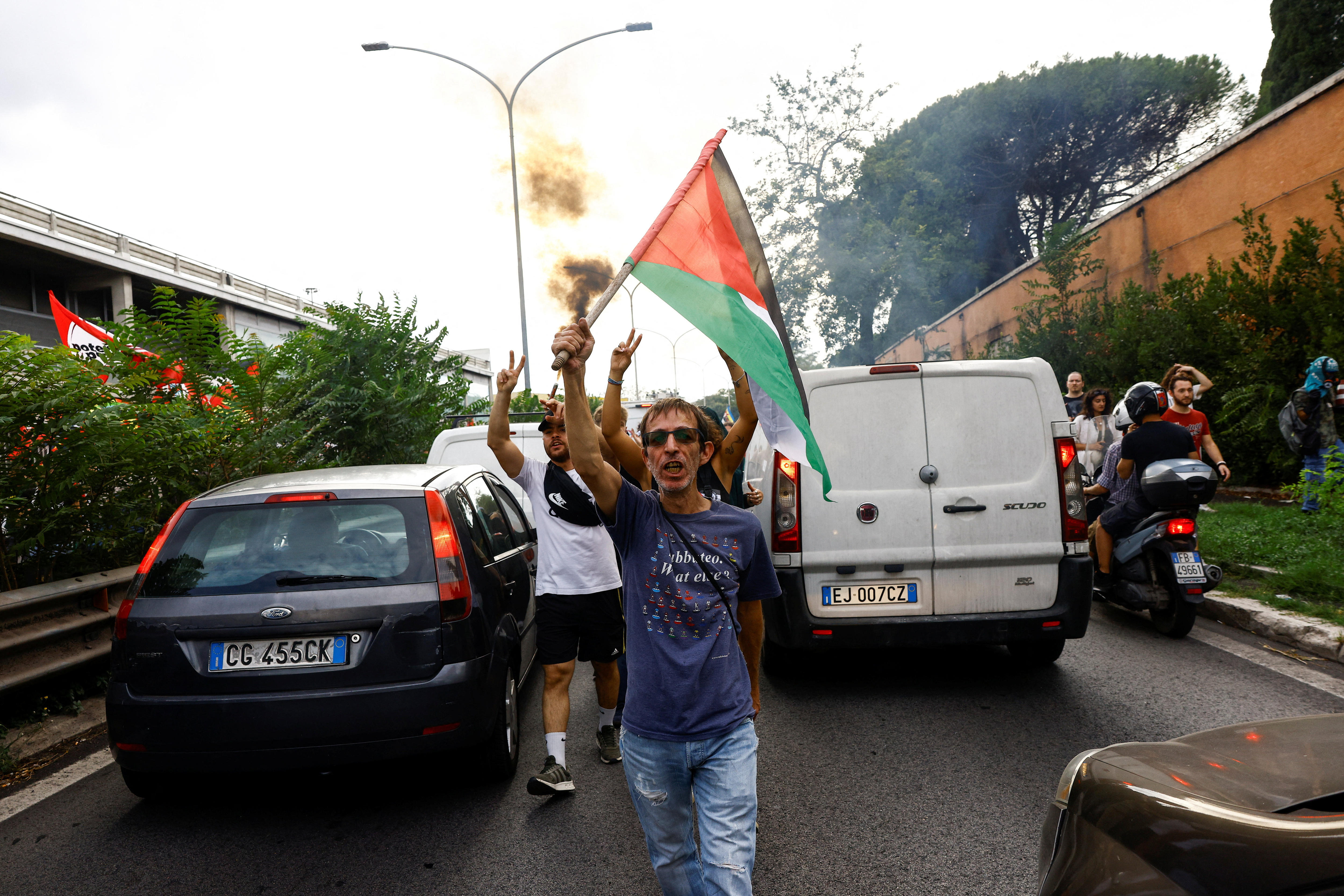 A group of men walking down the centre of a two-lane highway holding Palestinian flags, with stopped traffic around them.