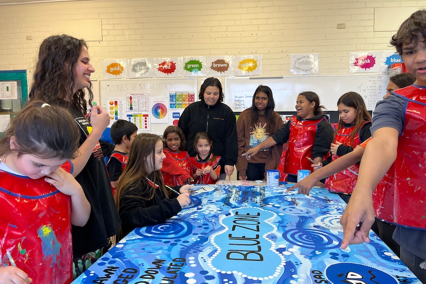 A woman and a group of children with paint brushes gather round a large mural on a table in tones of blue.