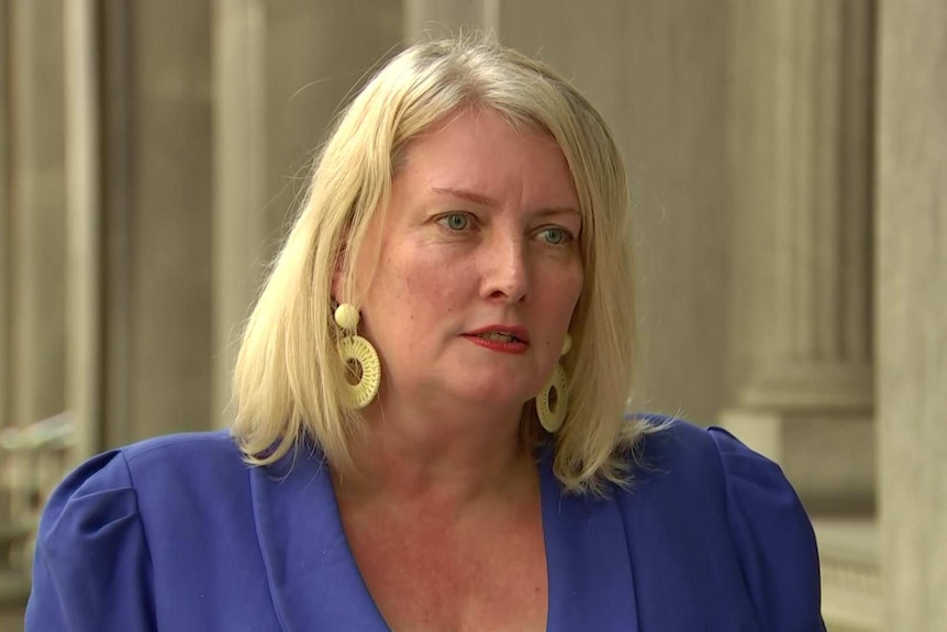 A blonde woman wearing a blue jacket in front of columns