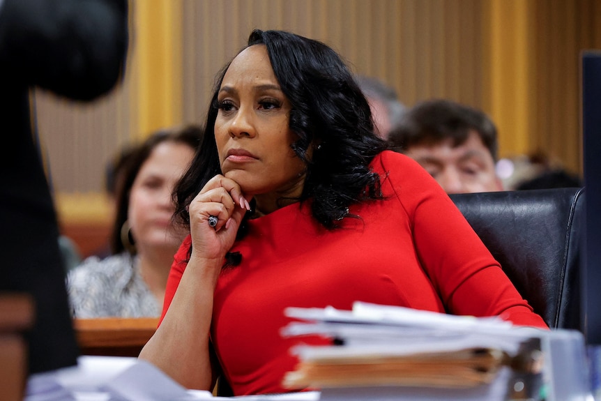 A middle-aged woman in a red outfit looks serious as she sits at a table in court.