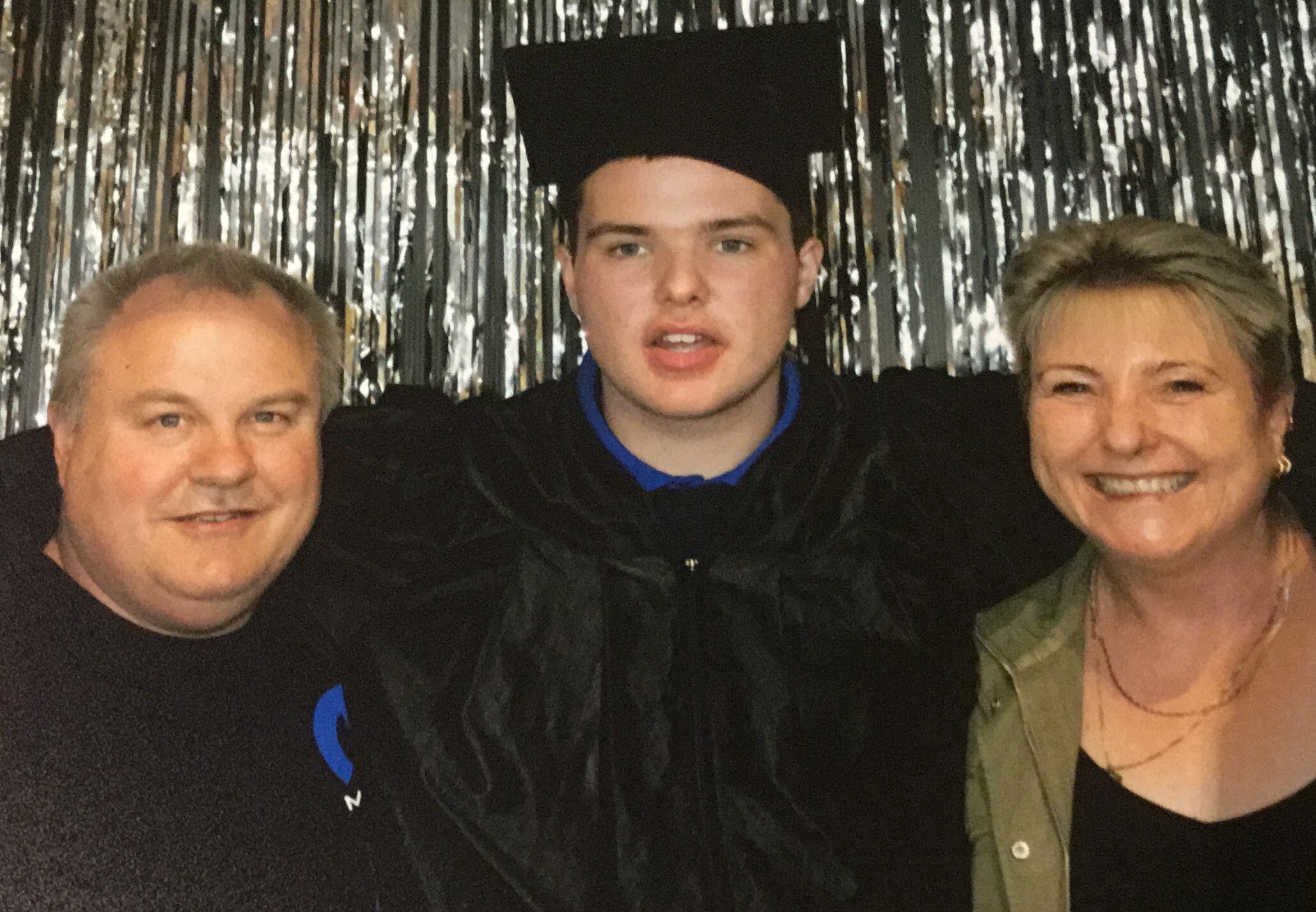 Boy in graduation gown and cap with arms around man and smiling woman.