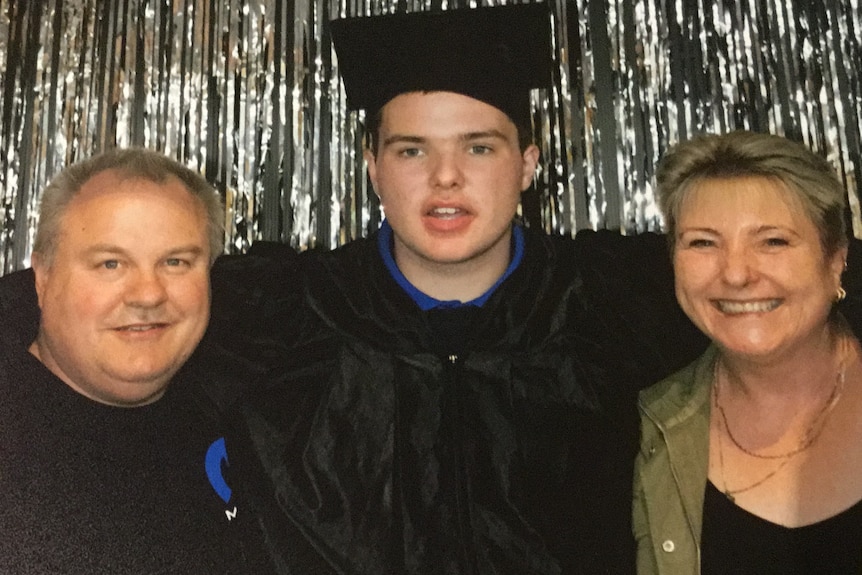 Boy in graduation gown and cap with arms around man and smiling woman.