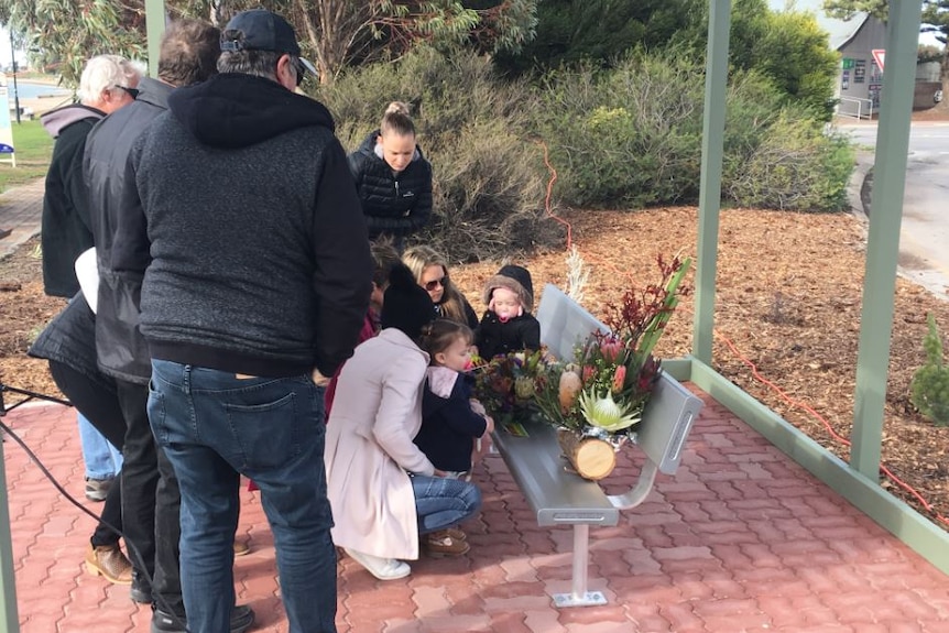 A group of people stand beside a bench and place flowers in memory of a loved one