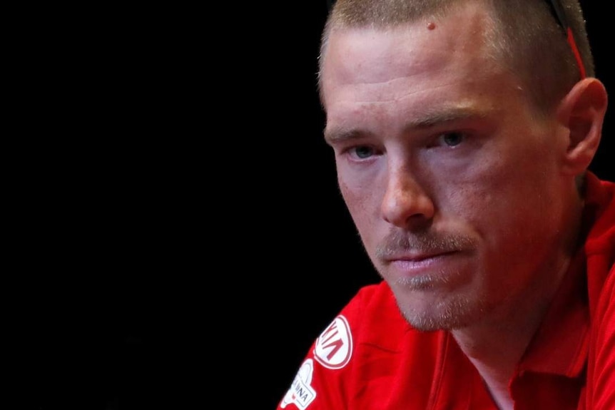 Two men in red cycling team polo shirts look down behind microphones in while they sit in front of a pitch black backdrop.