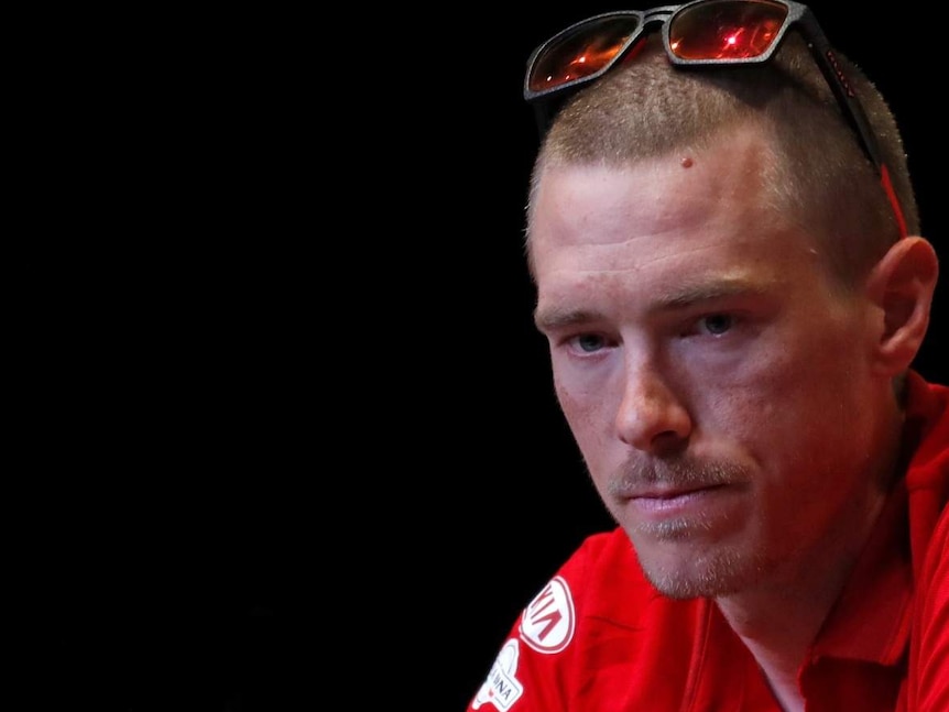 Two men in red cycling team polo shirts look down behind microphones in while they sit in front of a pitch black backdrop.