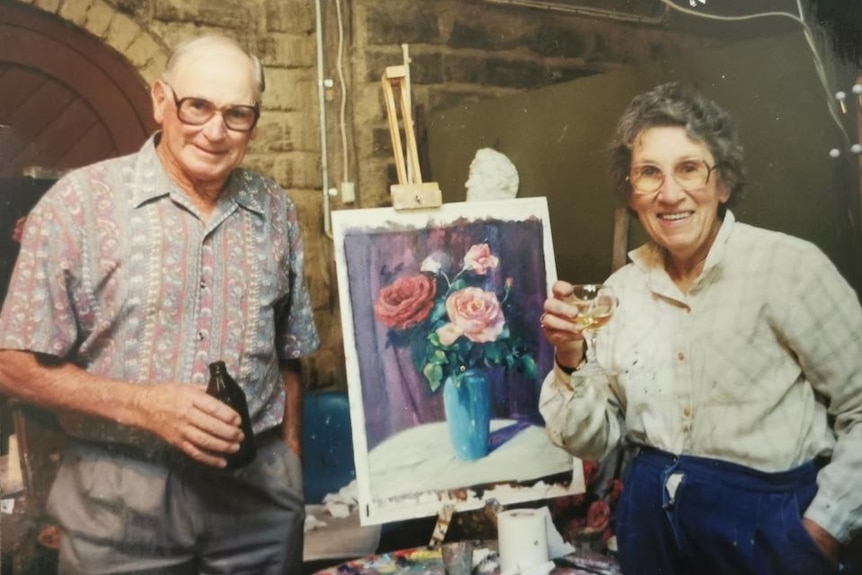 A middle-aged man and woman smiling and standing beside a painting.