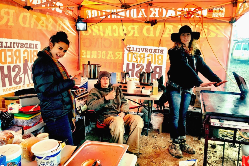 Two women stand in a tent , a man is seated. Inside the tent is cooking aparatus. 