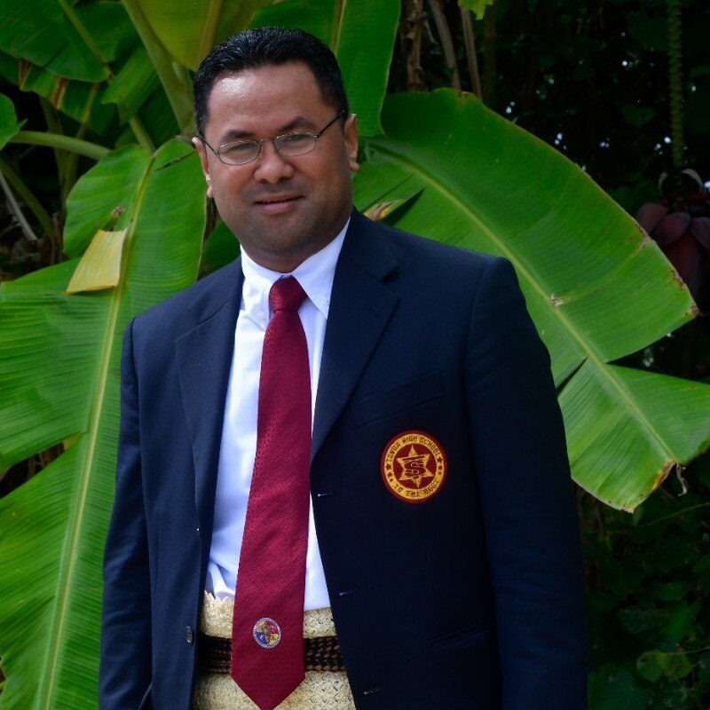 A Tongan man wearing glasses and a navy blue suit standing in front of green banana leaves