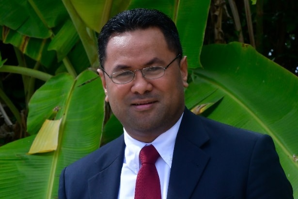 A Tongan man wearing glasses and a navy blue suit standing in front of green banana leaves