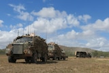 Army tanks driving in a paddock under a blue sky.