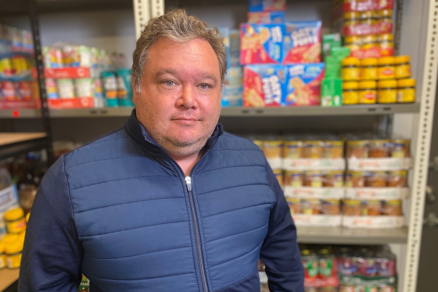 A man in a puffer jacket looks at the camera as he stands in front of shelves stacked with tinned food.