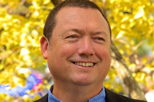 A man wearing a check shirt and dark jacket smiles, with autumn trees in the background.