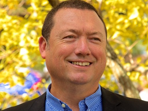 A man wearing a check shirt and dark jacket smiles, with autumn trees in the background.