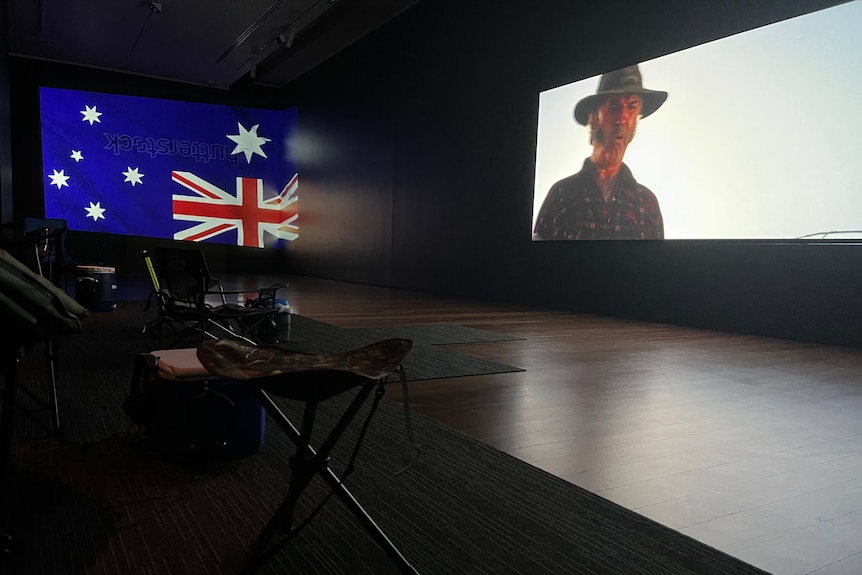 Two screens hang in a dark gallery space, one on each wall. On one is Wolf Creek, on the other an upside-down Australian flag.