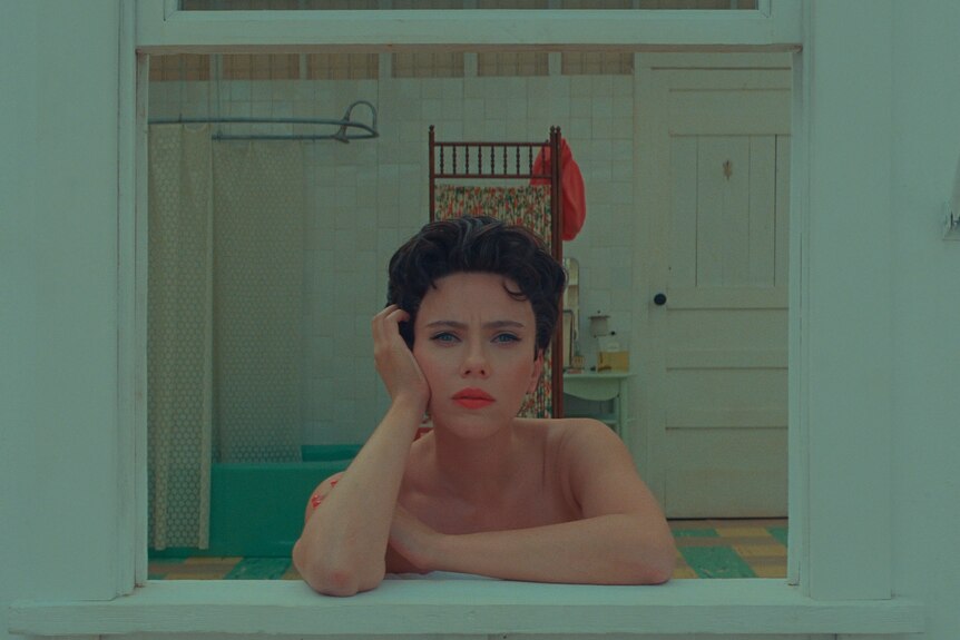 A white woman with short brunette hair wears red lipstick and leans out of the bathroom window of a small house in the desert.