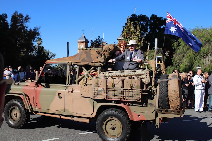 A woman and an elderly man wearing war medals ride in the back of an army truck in the parade.