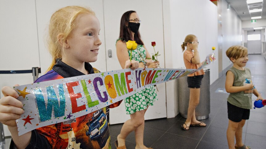 A woman and her children gather at the Darwin Airport, holding a sign that says "welcome home".