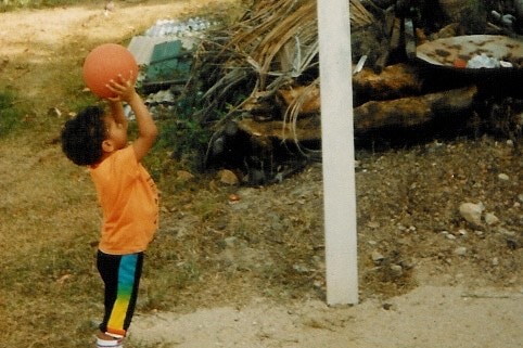 A little boy, about two years old, wearing an orange shirt, is shooting a basketball