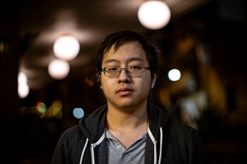 A man with short dark hair, wire frame glasses stands under glowing lights.