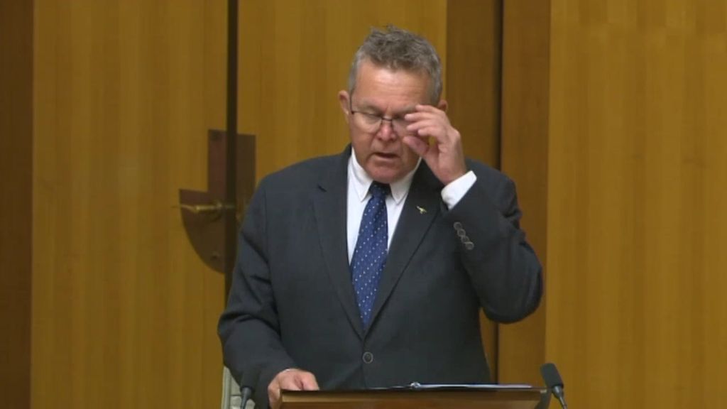 A man in a suit and tie adjusts his glasses while speaking at a lectern.