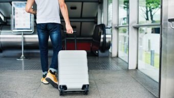 A young man wheels his carry on luggage through an airport, towards an escalator.