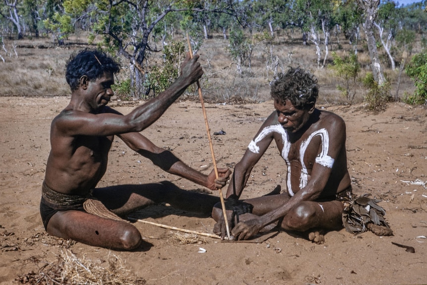 Two Indigenous men, one adorned with white paint, sit in the dirt making a fire with fire sticks.