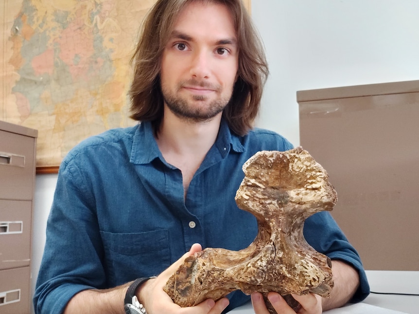 A young man with long, neat dark hair sits in a lab holding a partial skull.