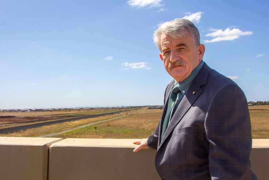 An older man stands in front of a train track in the distance