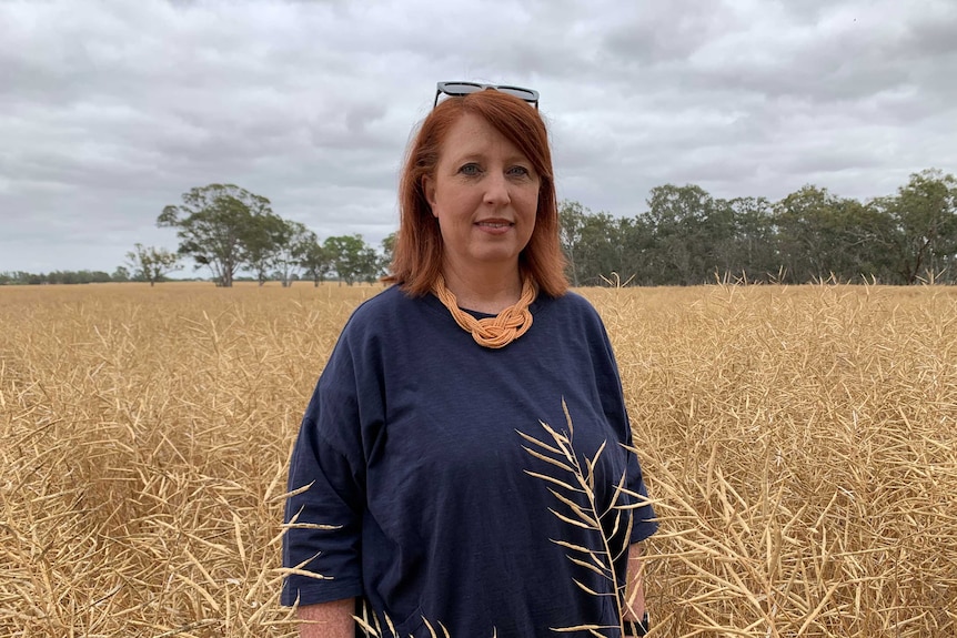 A woman with red hair, blue shirt and orange necklace stands in a canola crop that's ready to harvest.