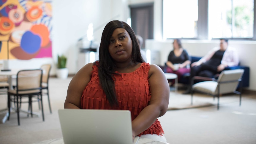 A woman looking pensive in front of her laptop, an office and colleagues in the background, she's recently been made redundant.