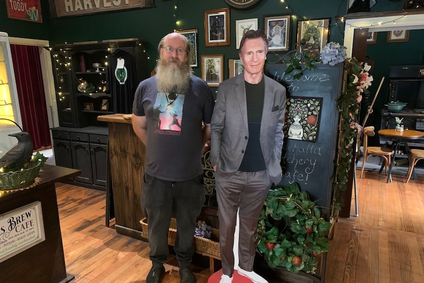 A balding man with long white beard standing next to a cutout of Liam Neeson