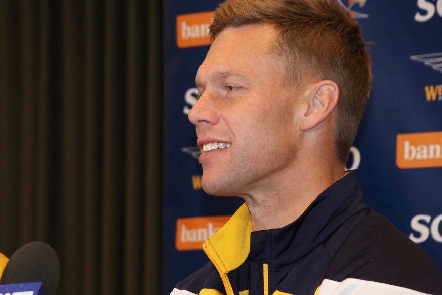 A side-on head and shoulders shot of a smiling Sam Mitchell indoors at a media conference.