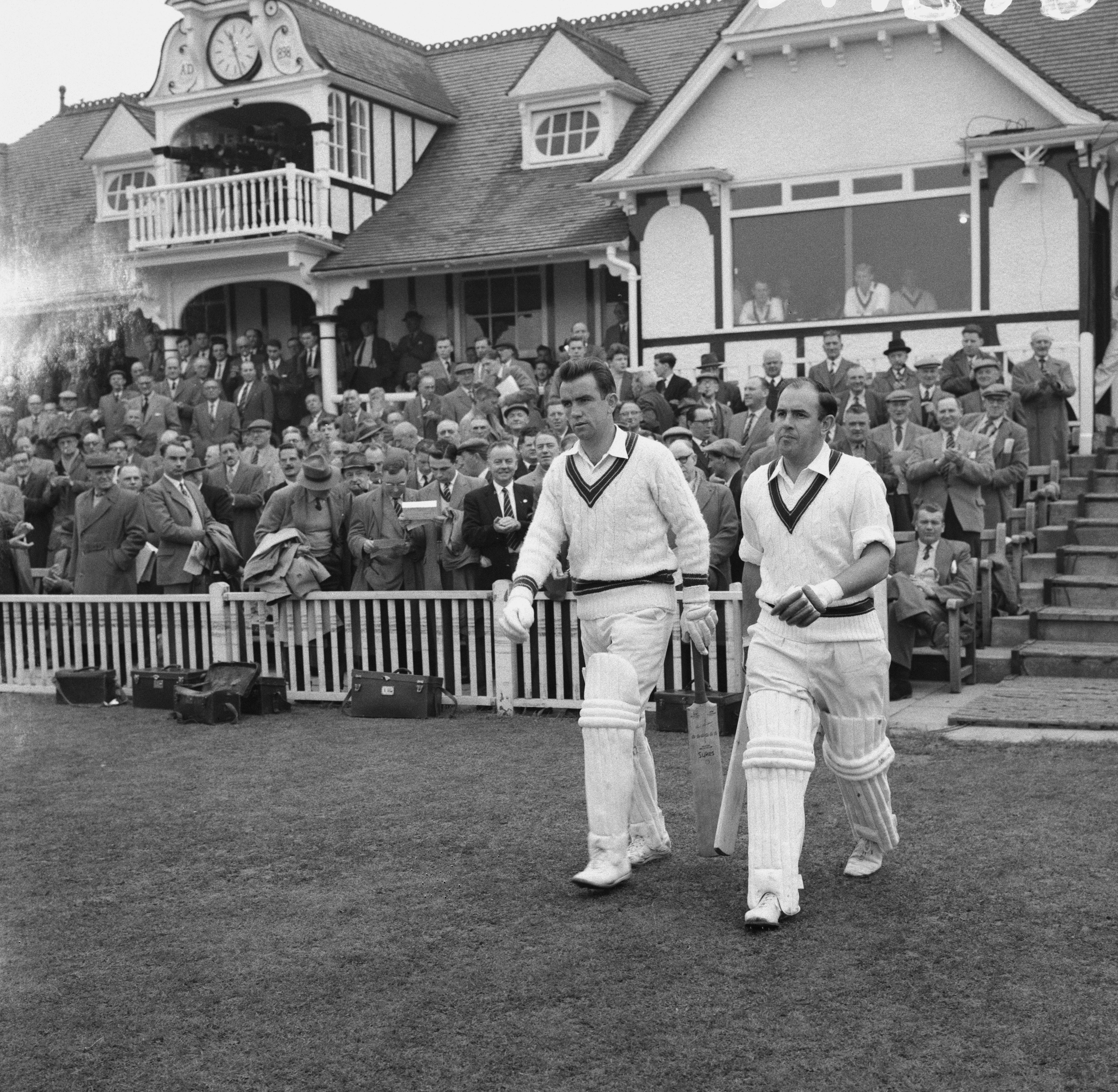 Two Australian cricketers in their whites walk out to bat as they are watched from the stands behind them by fans in the 1960s.