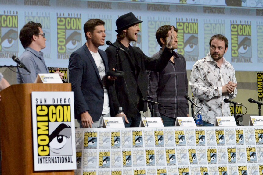 Five men stand in line behind table with namecards and comic-con logos. 