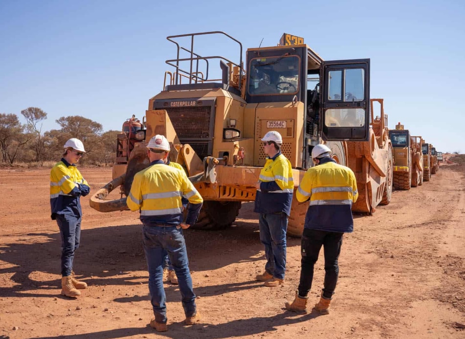 Four people in high-vis construction gear in front of heavy machinery.